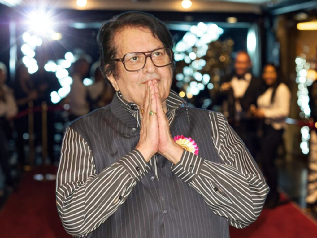 "Veteran Bollywood actor Manoj Kumar greets the audience with folded hands at an award ceremony, wearing a black vest over a striped shirt."