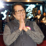 "Veteran Bollywood actor Manoj Kumar greets the audience with folded hands at an award ceremony, wearing a black vest over a striped shirt."