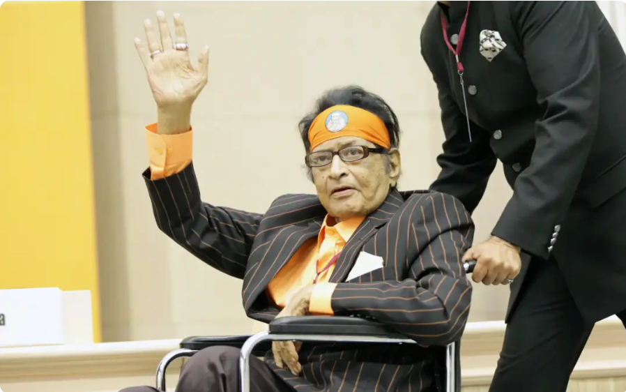 "Legendary Bollywood actor Manoj Kumar in a wheelchair, waving to the crowd during a public appearance, wearing an orange headband and a pinstripe suit."